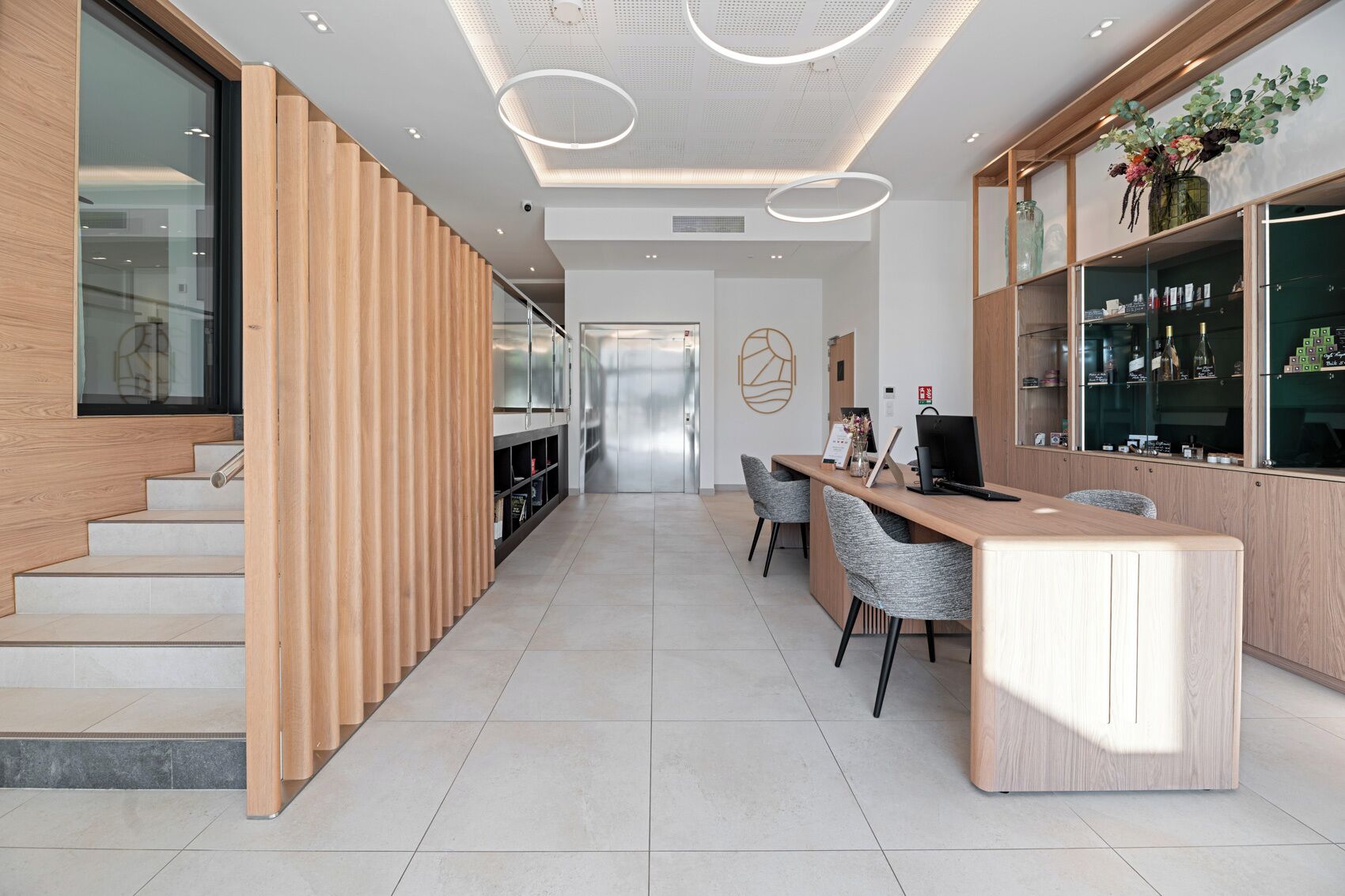 Een moderne hotel lobby met een houten receptiebalie, grijze stoelen, een trap en een glazen wand.