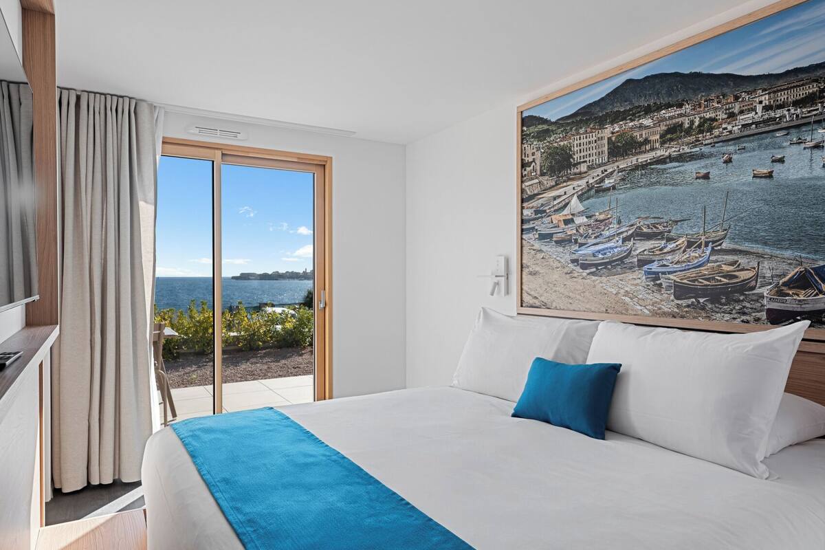 A hotel room with a bed, a TV, a painting of a coastal scene, and a view of the sea through a sliding door.
