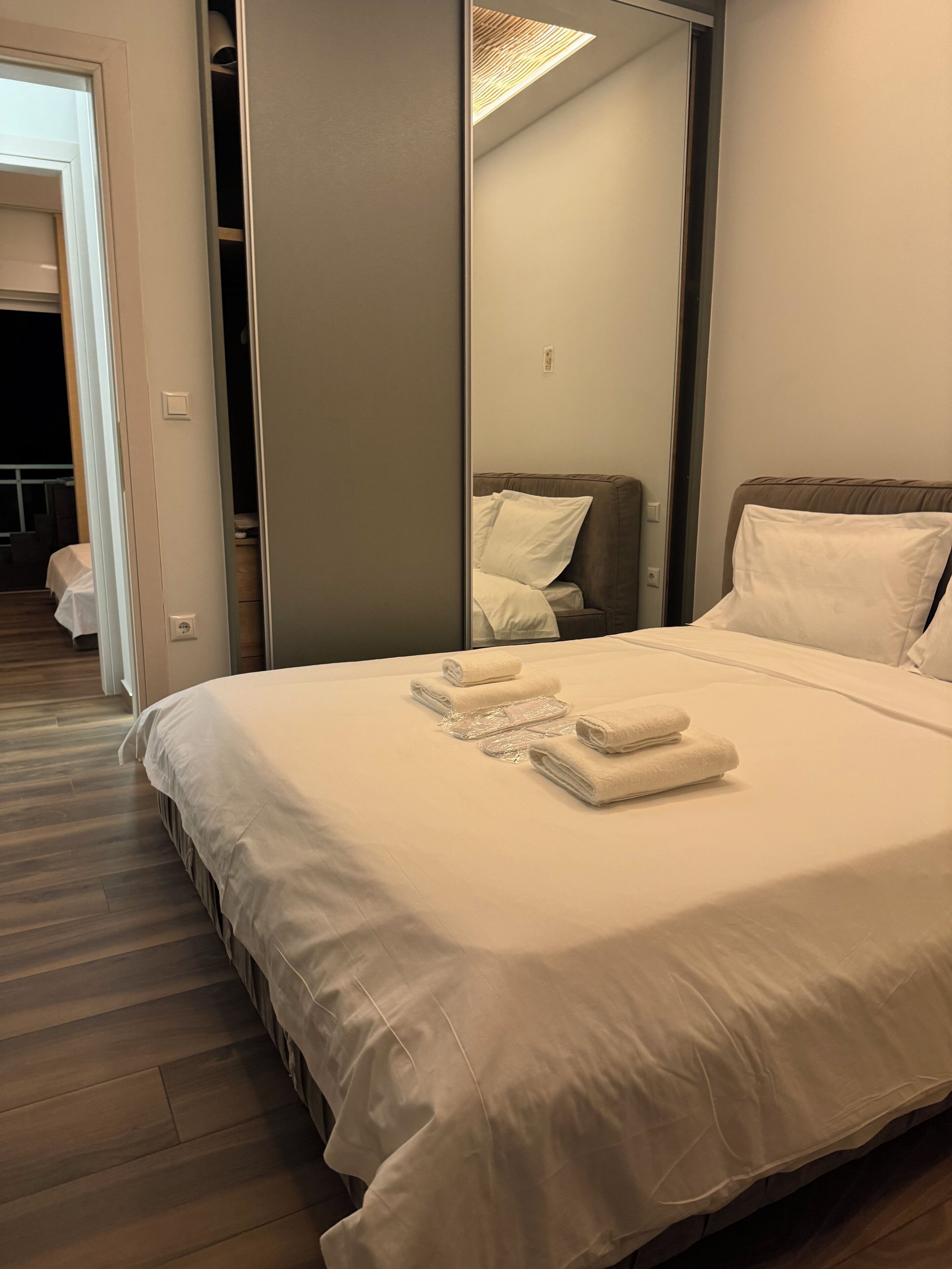 Una cama bien hecha con sábanas y toallas blancas, un cabecero marrón y un espejo grande que refleja la habitación.