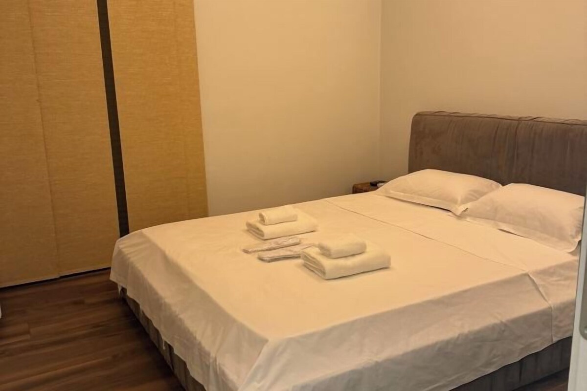 A neatly made bed with white linens and towels, a wooden floor, and a plain wall.