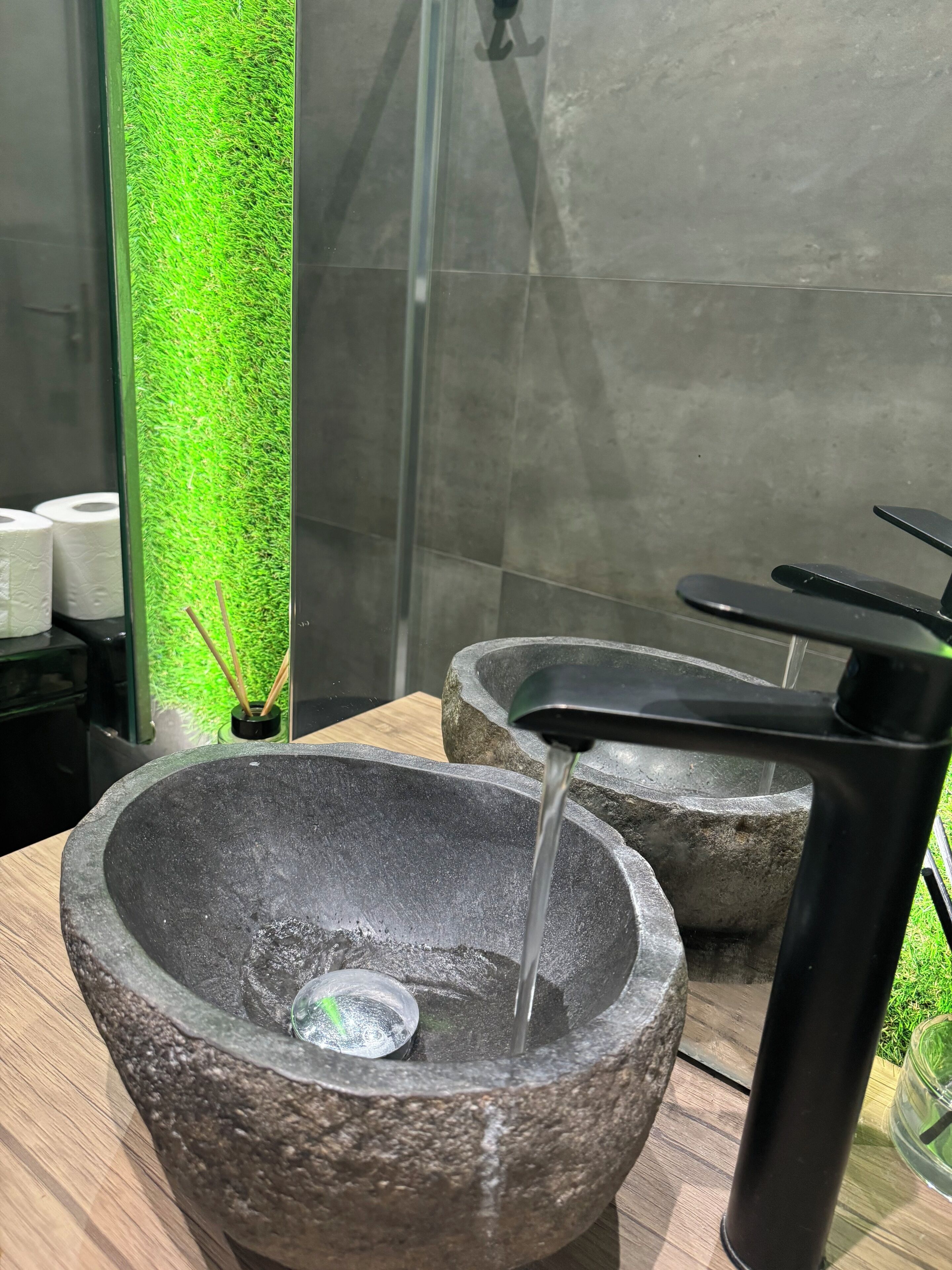 Ein Steingefäß mit Wasser, das aus einem modernen Wasserhahn fließt, ein Glasbehälter mit einer grünen Pflanze und eine Holzoberfläche.