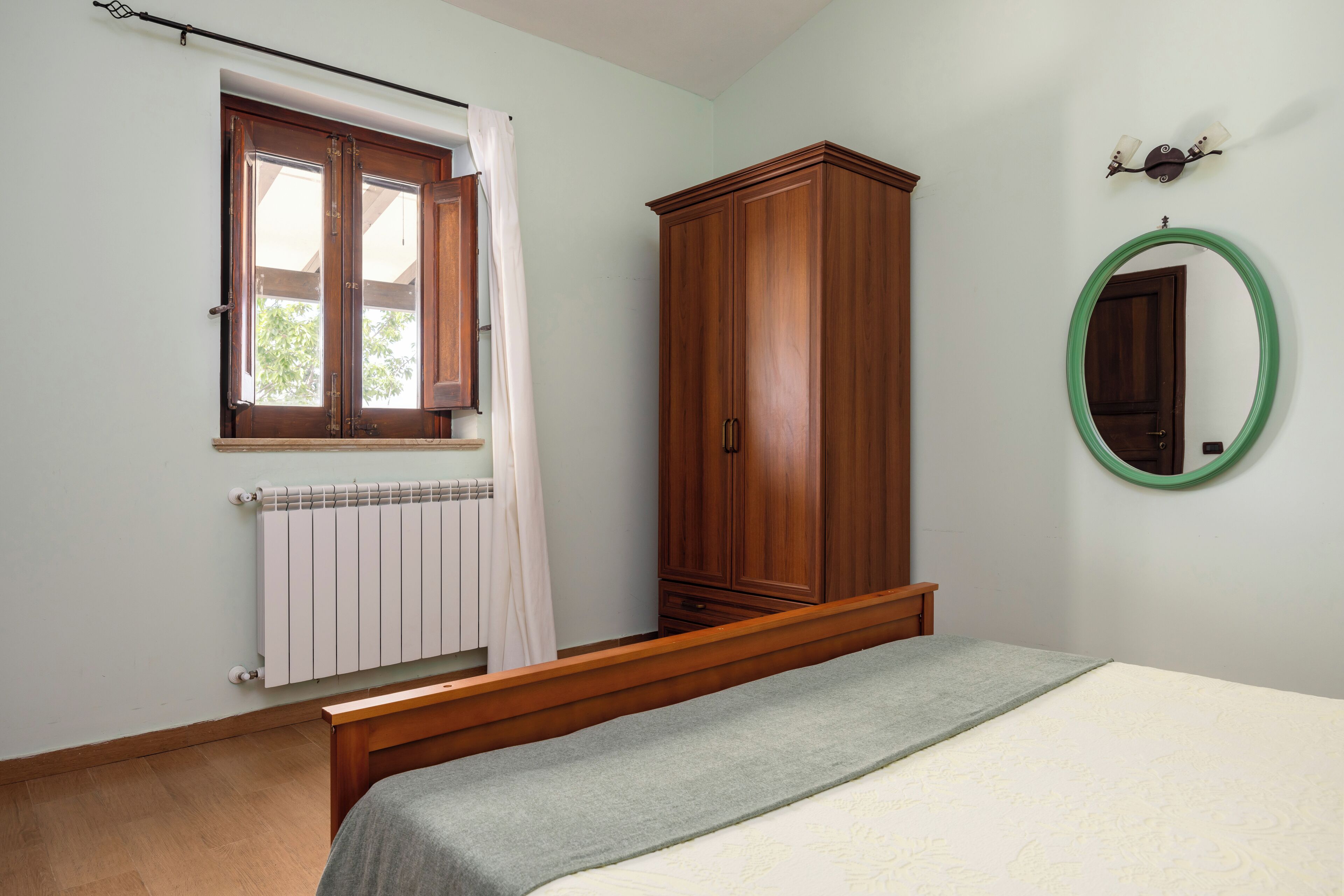 A bedroom with a wooden bed, a radiator, a wardrobe, and a window with a curtain.