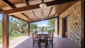 An outdoor covered patio with a stone wall and wooden beams, a table with chairs, and a view of trees and water. - Villa Granci con Piscina by Wonderful Italy (Polizzi Generosa)