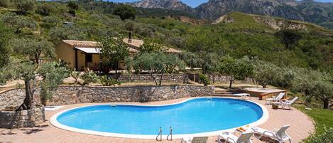 A swimming pool with lounge chairs, surrounded by a stone wall and greenery, with mountains in the background.