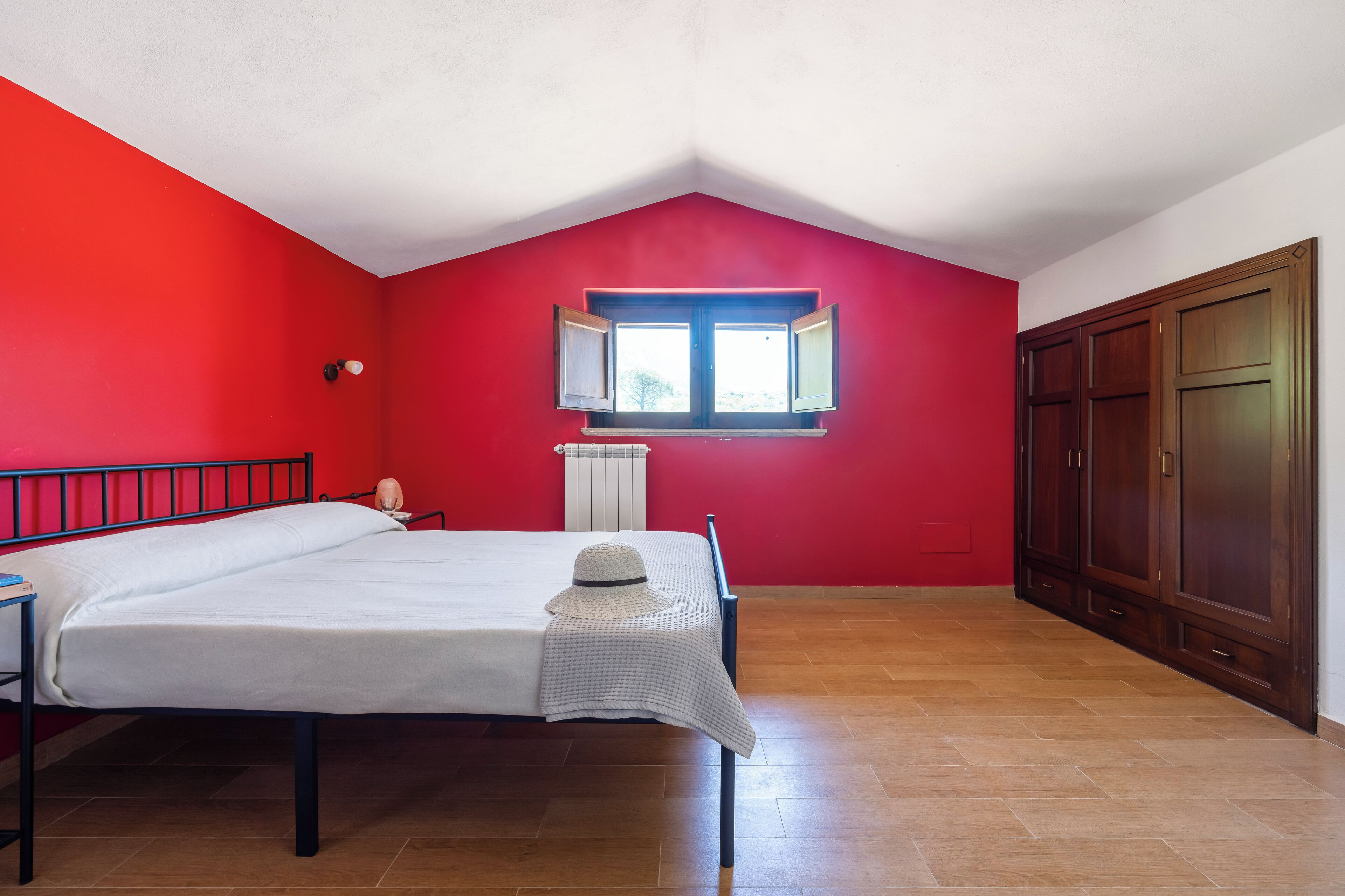 A bedroom with a red wall, a window, a bed, a wardrobe, and a radiator.