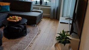 A modern living room with a sofa, coffee table with fruit, a small ottoman, a potted plant, and a TV. - Apartment Busch in the center (Bad Harzburg)