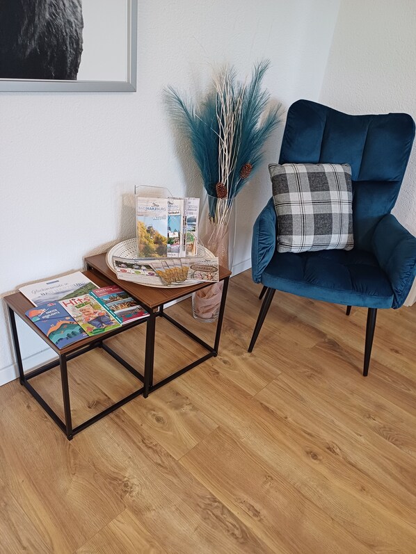 Una habitación con un sillón azul, una mesita de madera con revistas y un jarrón decorativo con plumas azules.