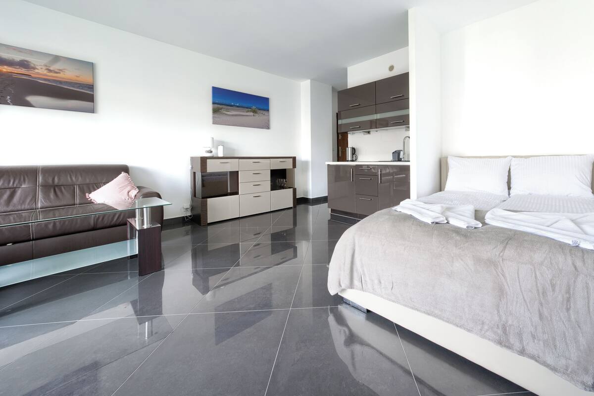 A modern hotel room with a large bed, a sofa, a kitchenette, and a view of the beach.