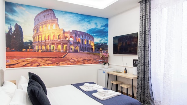 Habitación de hotel con un cuadro grande del Coliseo, una cama con ropa blanca, un escritorio con una computadora y un refrigerador pequeño.
