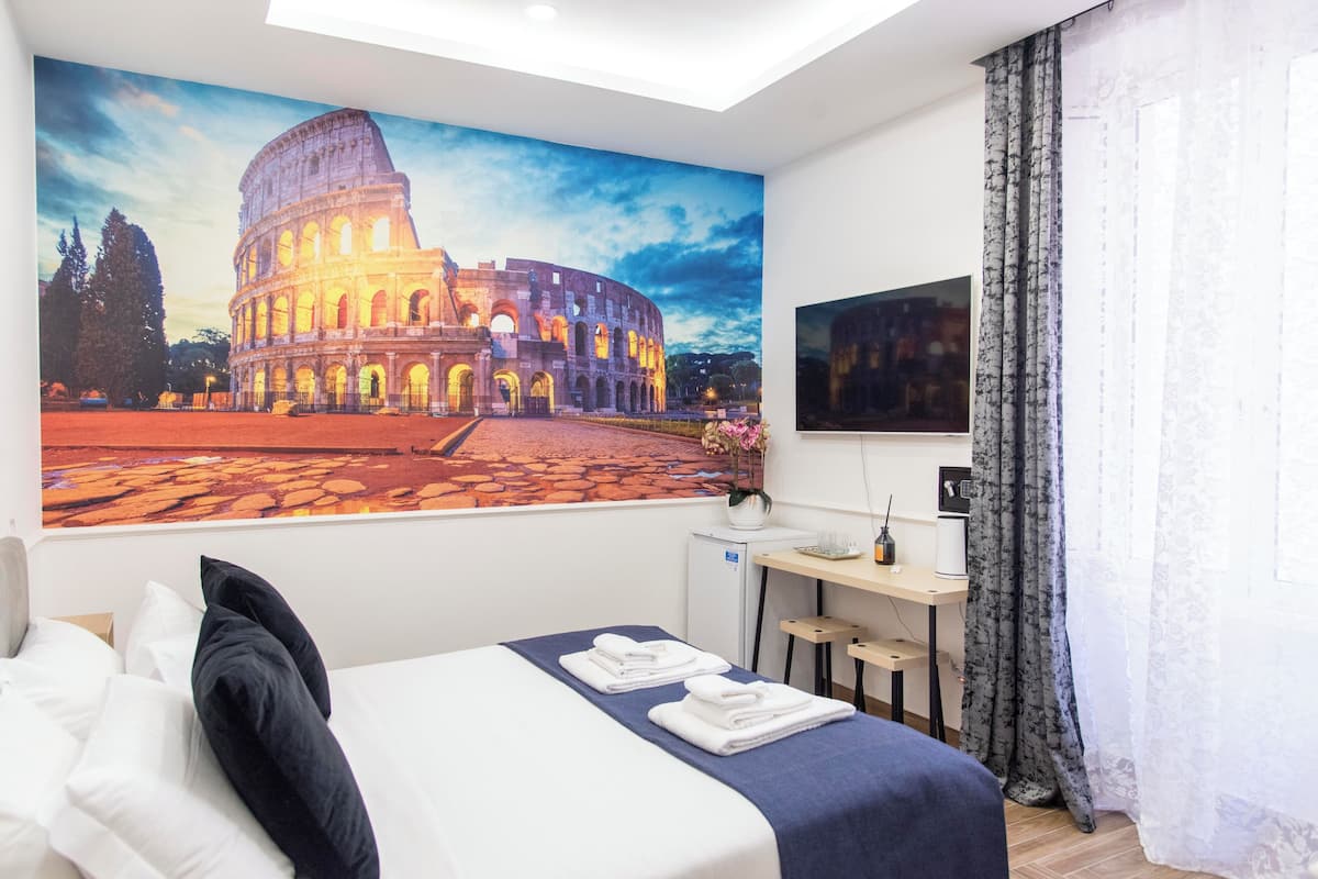 A hotel room with a large painting of the Colosseum, a bed with white linens, a desk with a computer, and a small refrigerator.