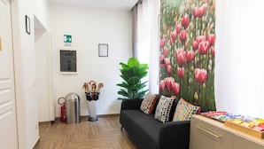 Un vestíbulo de hotel con un sofá negro, cojines de colores, una gran planta en maceta, un extintor y un letrero de salida de emergencia instalado en la pared.
