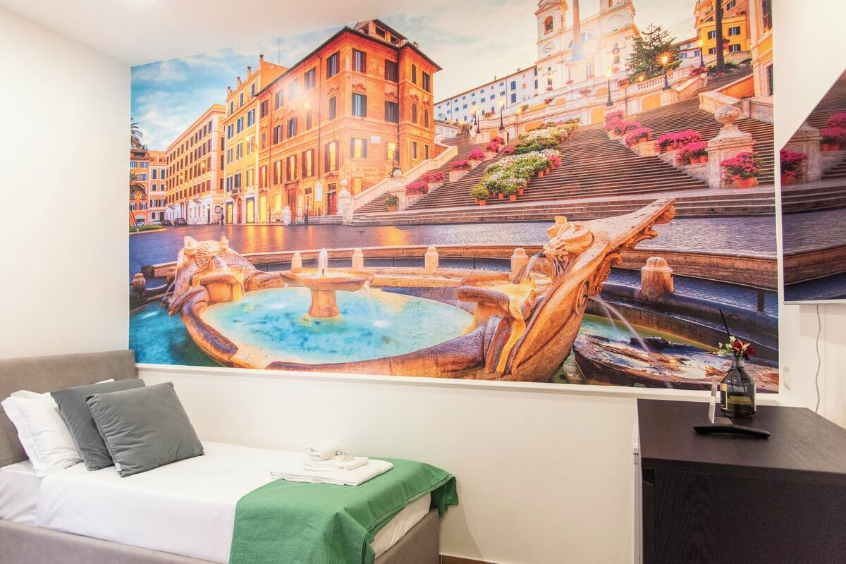 A hotel room with a large mural of a historic cityscape, a bed with a green throw, and a desk with a vase.