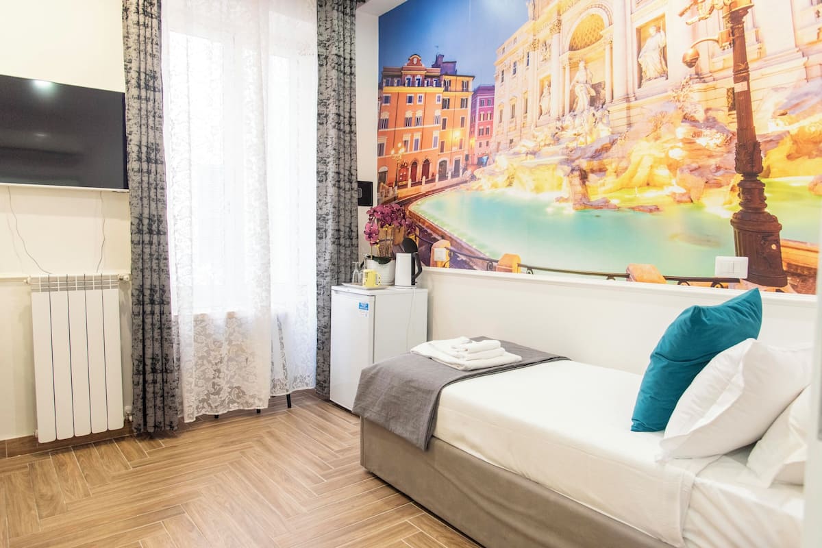 A hotel room with a bed, a small desk, a radiator, a TV, and a mural of a cityscape.
