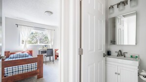 Un dormitorio con cama de madera, un escritorio y una silla. Hay un baño con lavabo y espejo.