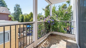 Un balcón cubierto con barandilla de metal, suelo de piedra y vista a un jardín con flores moradas.