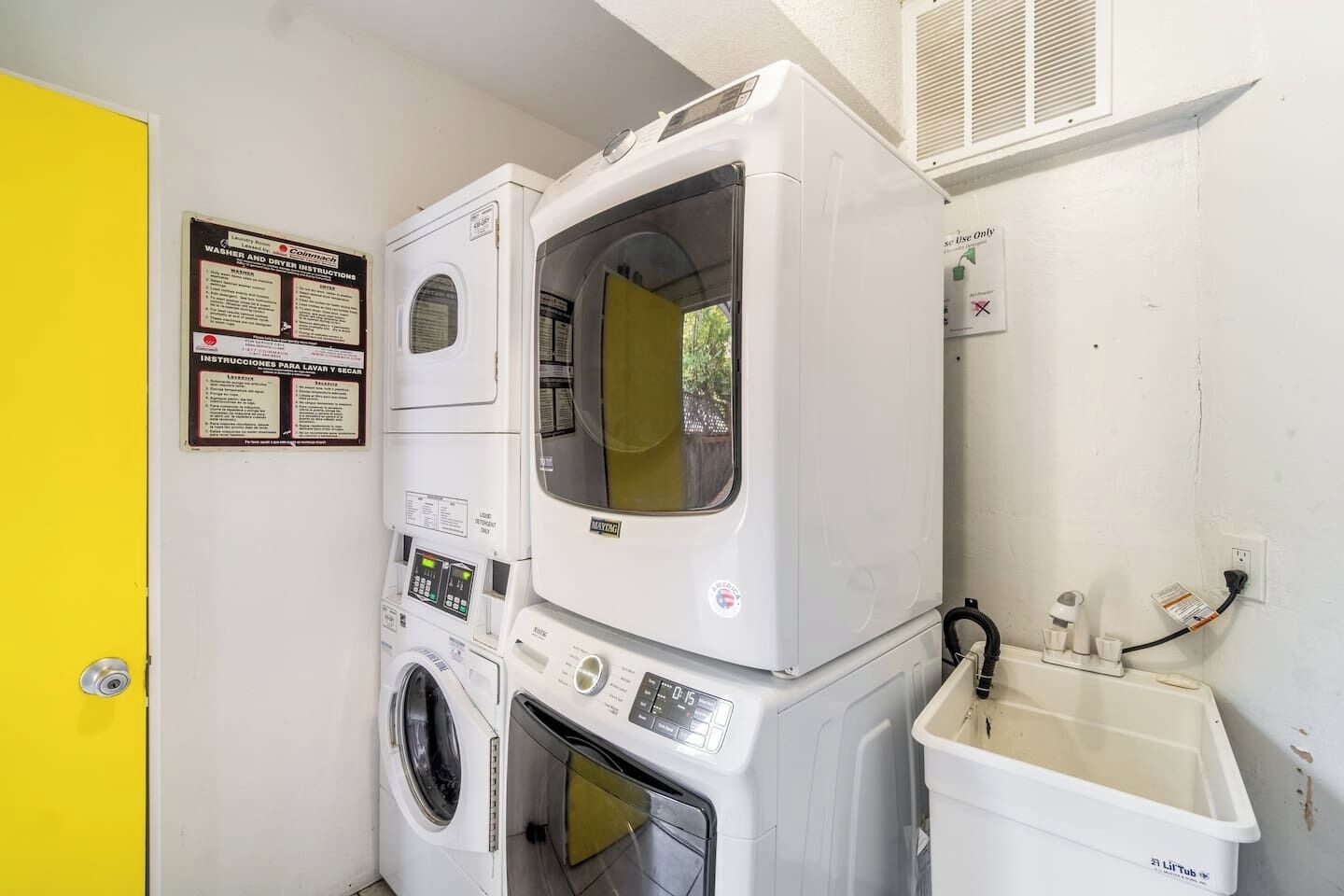 A laundry area with stacked washers and dryers, a washing machine, a drying machine, and a small sink with a hose. Instructions for use are posted on the wall.