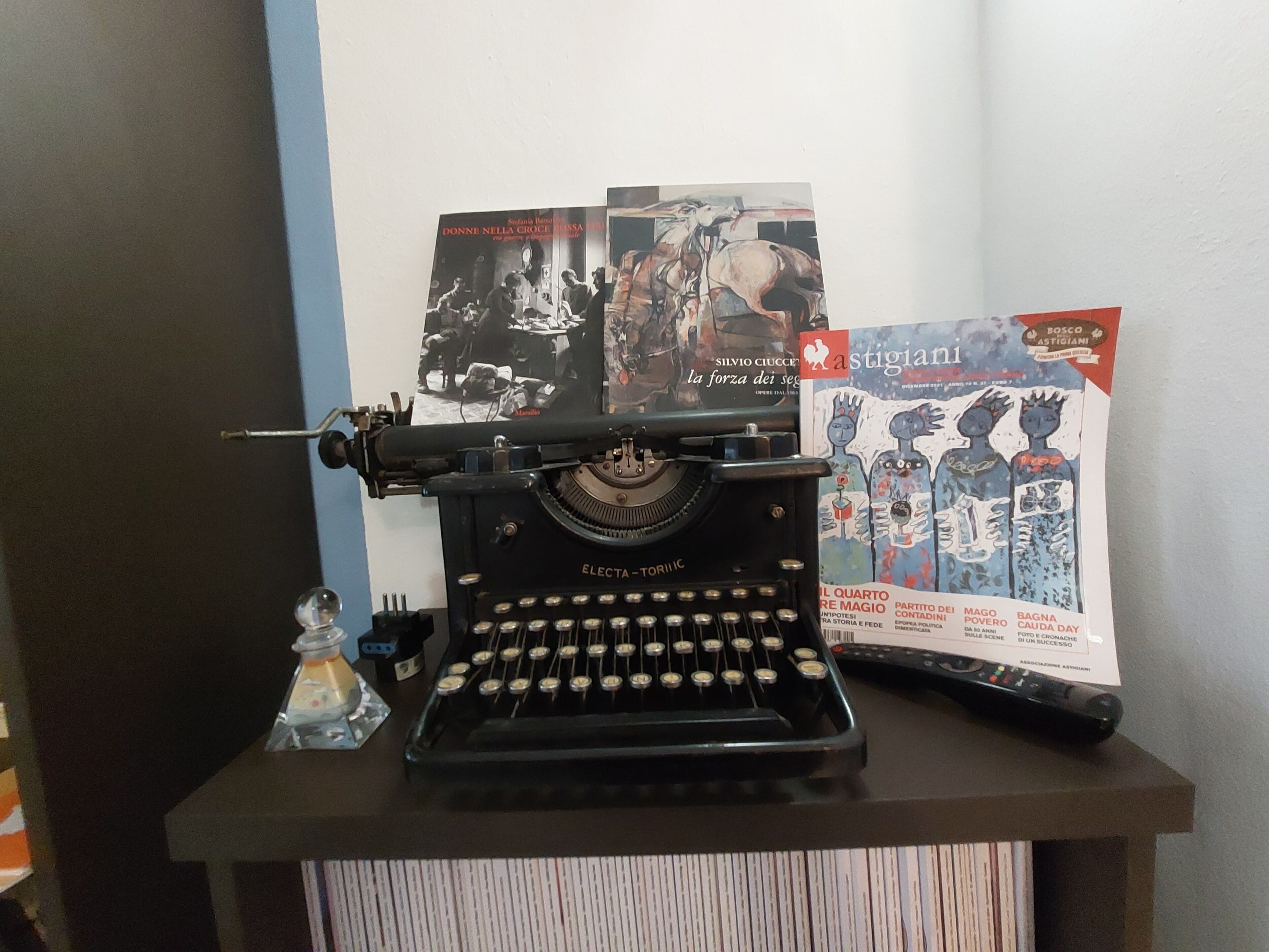 Uma máquina de escrever Electa antiga sobre uma mesa, com um livro e uma revista.