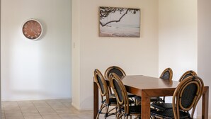 Un comedor con una mesa de madera y seis sillas de mimbre. Se ve un reloj de pared y un cuadro con motivos playeros.
