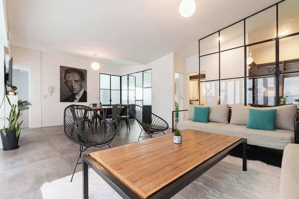 A modern living room with a sofa, coffee table, and chairs. There is a dining area with a table and chairs. A large framed portrait is on the wall.