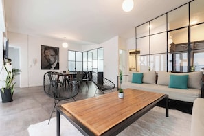 A modern living room with a wooden coffee table, black wire chairs, a sofa with teal cushions, and a large abstract painting on the wall.