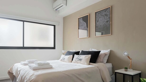 Een slaapkamer met een groot raam, een bed met witte lakens en kussens, een klein nachtkastje met een lamp, en twee ingelijste foto’s aan de muur.