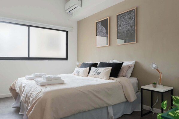 A bedroom with a large window, a bed with white linens and pillows, a small bedside table with a lamp, and two framed pictures on the wall.