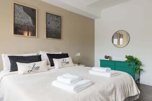 A hotel room with a bed, two framed maps on the wall, a bedside table with a lamp, a teal dresser, and a mirror.