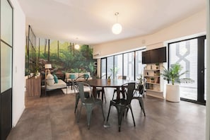 Un comedor amplio con una mesa grande y sillas, rodeado de ventanas grandes y un mural de una escena boscosa.