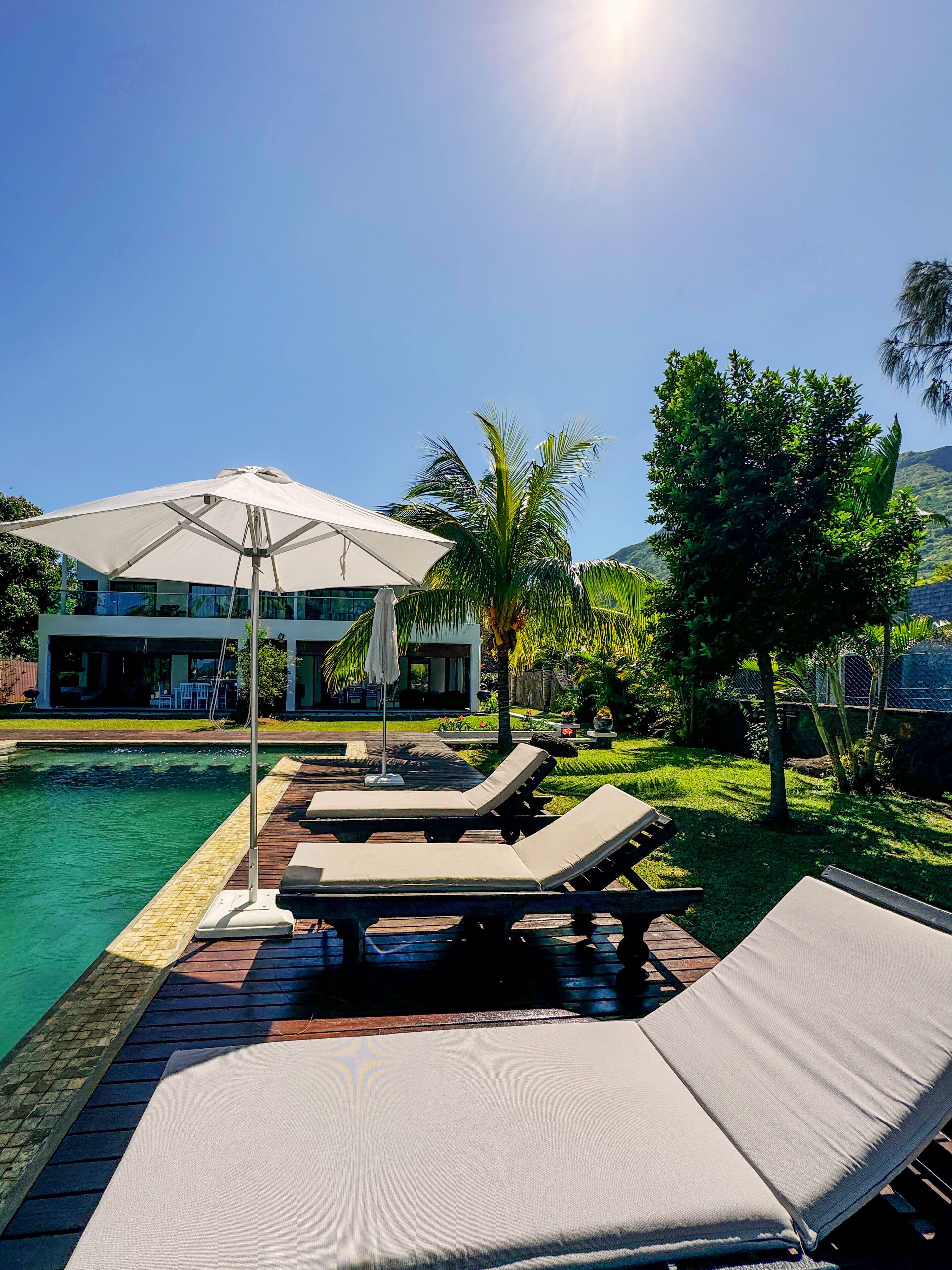 A poolside area with lounge chairs and a large umbrella, surrounded by lush greenery and a modern building in the background.
