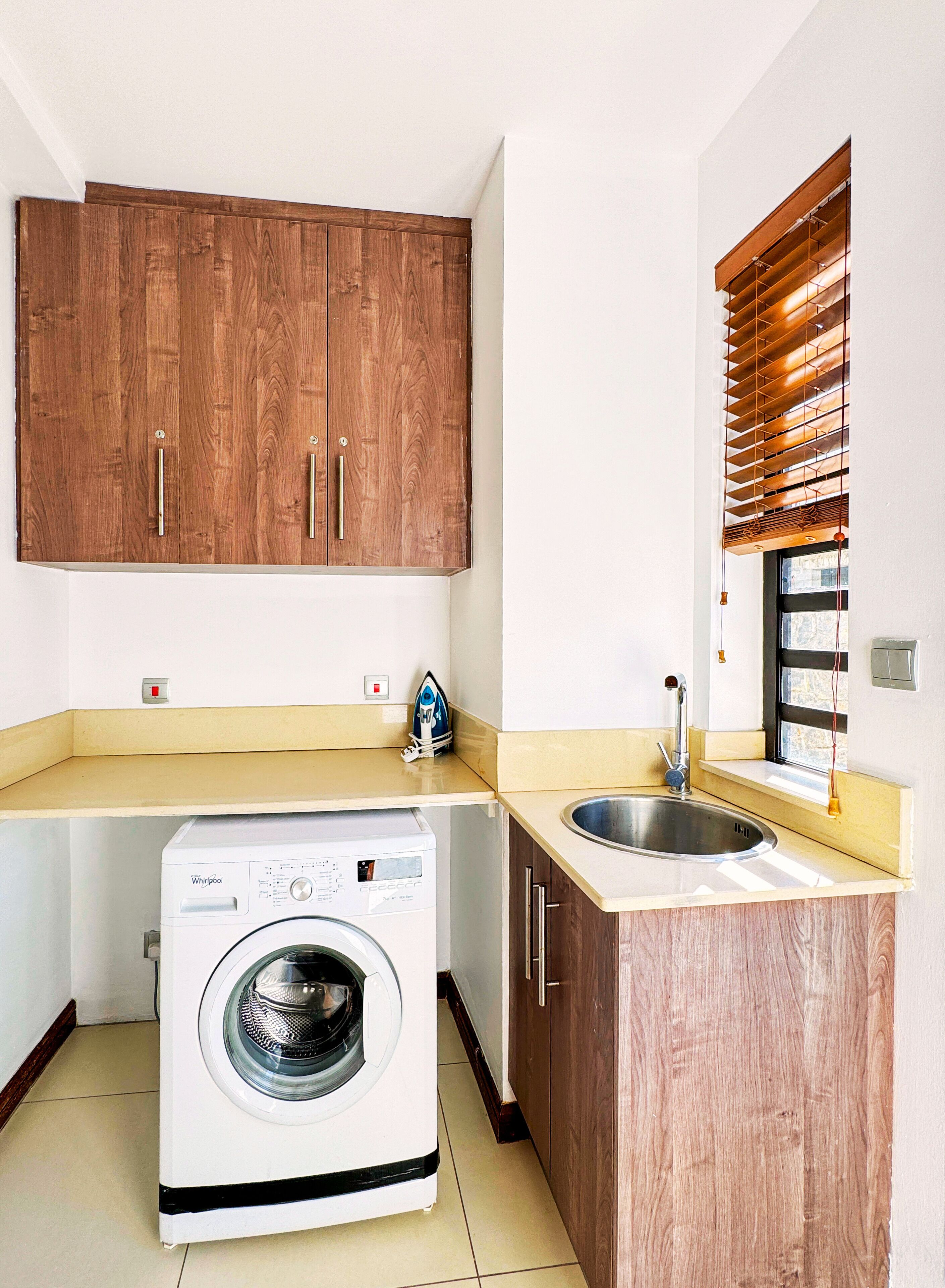 A compact kitchen with wooden cabinets, a built-in washing machine, a sink, and a window with blinds.