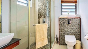 Ein Badezimmer mit Duschkabine aus Glas, Toilette, Waschbecken und einem Fenster mit Jalousien.
