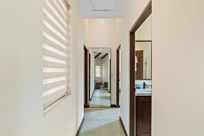 A narrow hallway with a ceiling vent, a window with blinds, and a door leading to a bathroom with a mirror and sink.