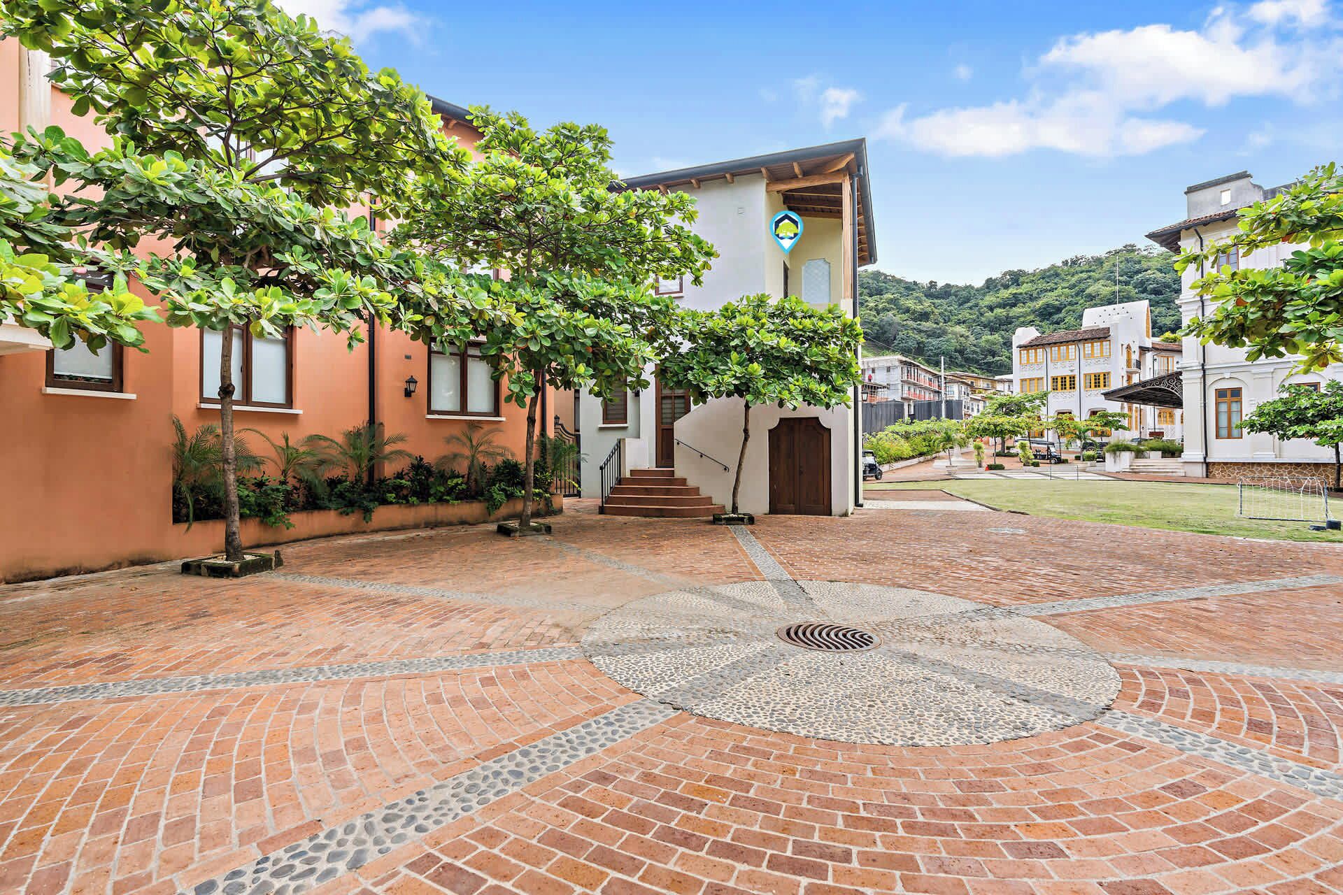A courtyard with a brick floor, a central circular mosaic, and a building with a white facade and a blue logo.
