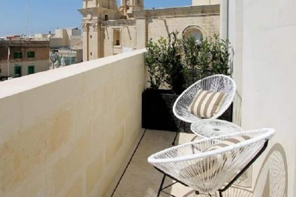 Een balkon met twee witte draadstoelen, een bloempot en uitzicht op twee historische gebouwen met klokkentorens.