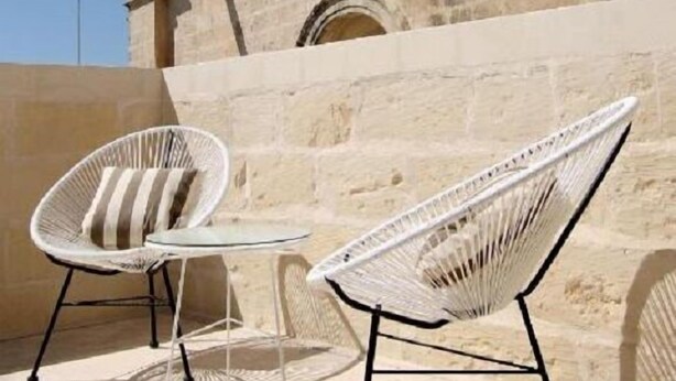 Two white wicker chairs with metal frames and a small round table, set against a stone wall with a clock tower in the background. - The King George Village Boutique Living (Hal Gharghur)