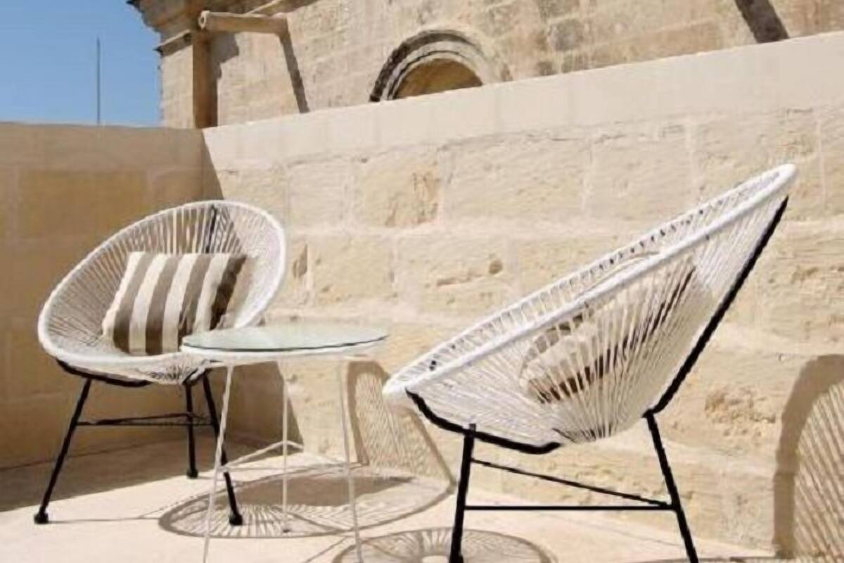 Twee witte rieten stoelen met metalen frames en een kleine ronde tafel, geplaatst tegen een stenen muur met een klokkentoren op de achtergrond.