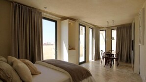 A hotel room with a bed, a dining table with chairs, and a view of the city through large windows. - The King George Village Boutique Living (Hal Gharghur)