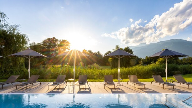 A poolside area with lounge chairs and umbrellas, surrounded by greenery and mountains in the background. - Dolomitengolf Suites (Lavant)