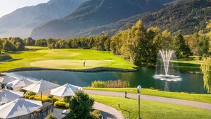 A golf course with a pond, surrounded by mountains and trees. - Dolomitengolf Suites (Lavant)