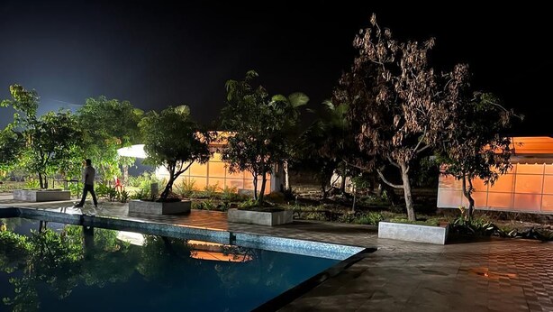 Un área de piscina nocturna con un deck cubierto de azulejos, rodeada de macetas y árboles.