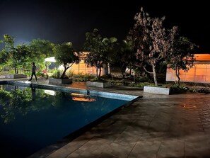 Un área de piscina nocturna con un deck cubierto de azulejos, rodeada de macetas y árboles.