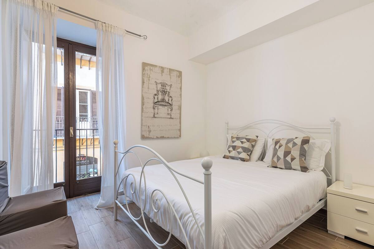 Camera da letto con un letto bianco, un comodino e vista su un balcone attraverso una porta con tende.