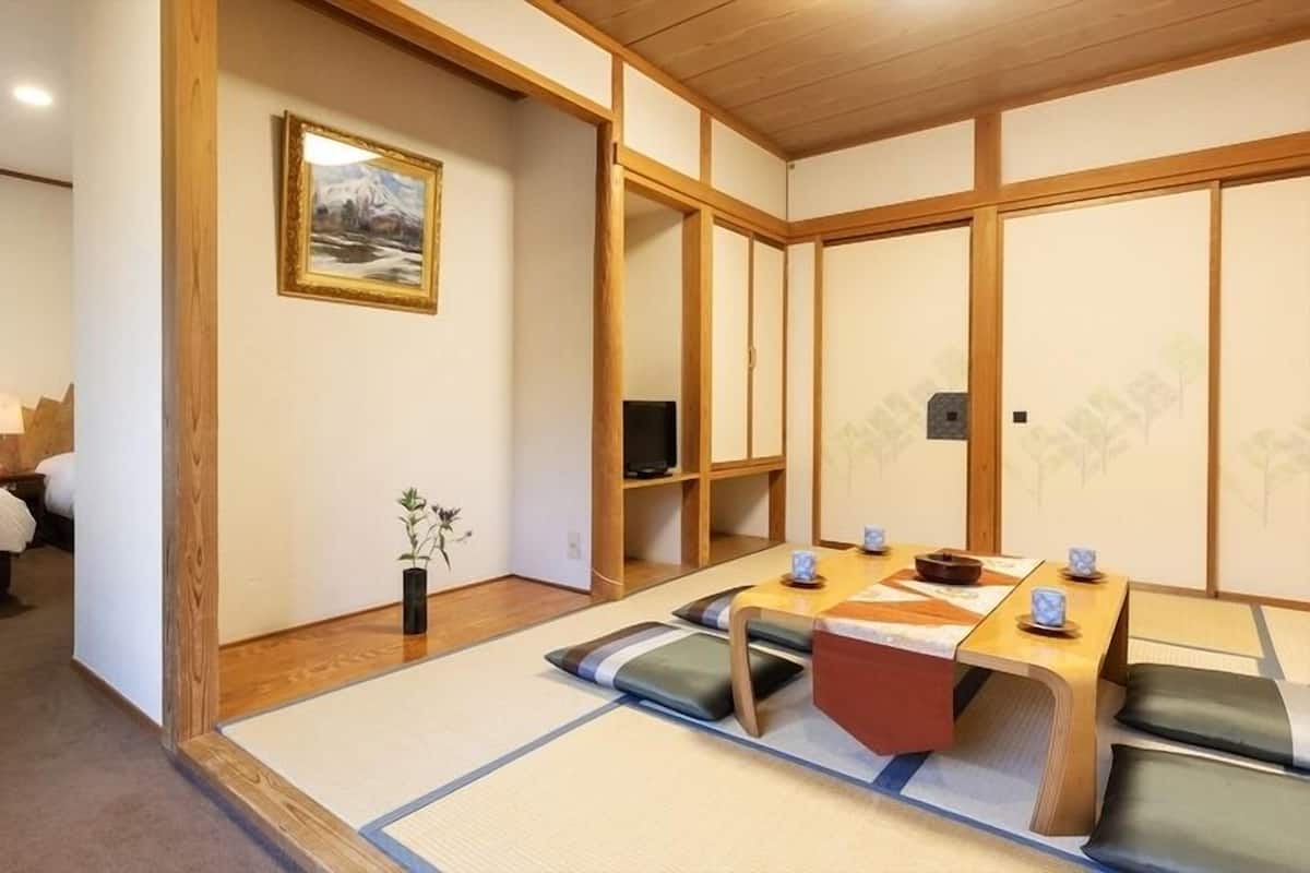 Una tradizionale stanza in stile giapponese con pavimento in tatami, un tavolino con cuscini, un letto con poggiatesta in legno e un quadro appeso al muro.