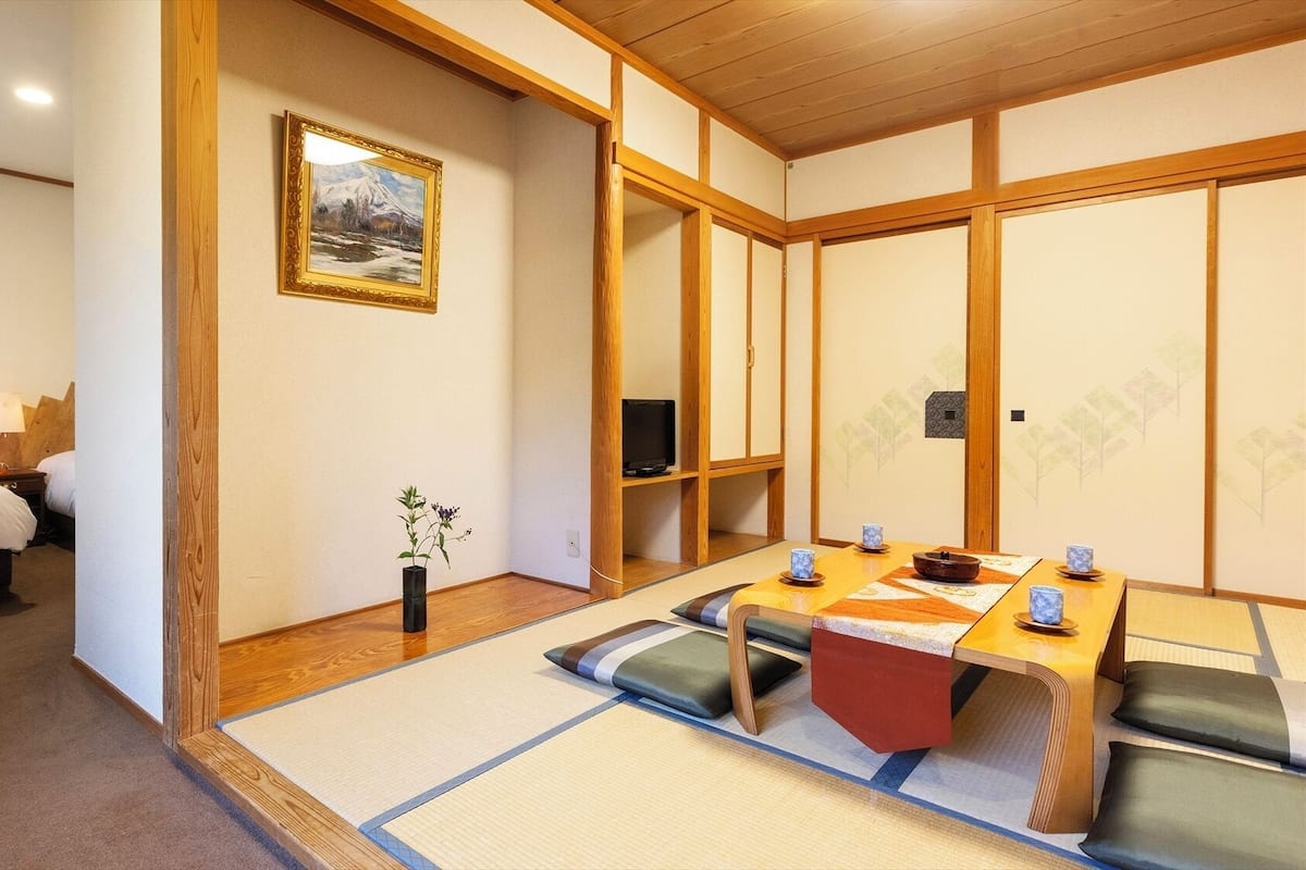 Una tradizionale stanza in stile giapponese con pavimento in tatami, un futon, una televisione e un tavolo da pranzo con cuscini.