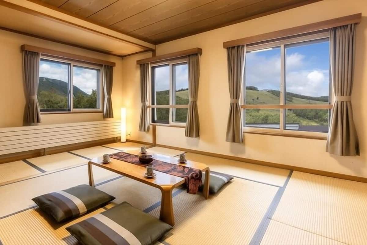 Una stanza con pavimento in tatami, un tavolino basso, cuscini e ampie finestre con tende.