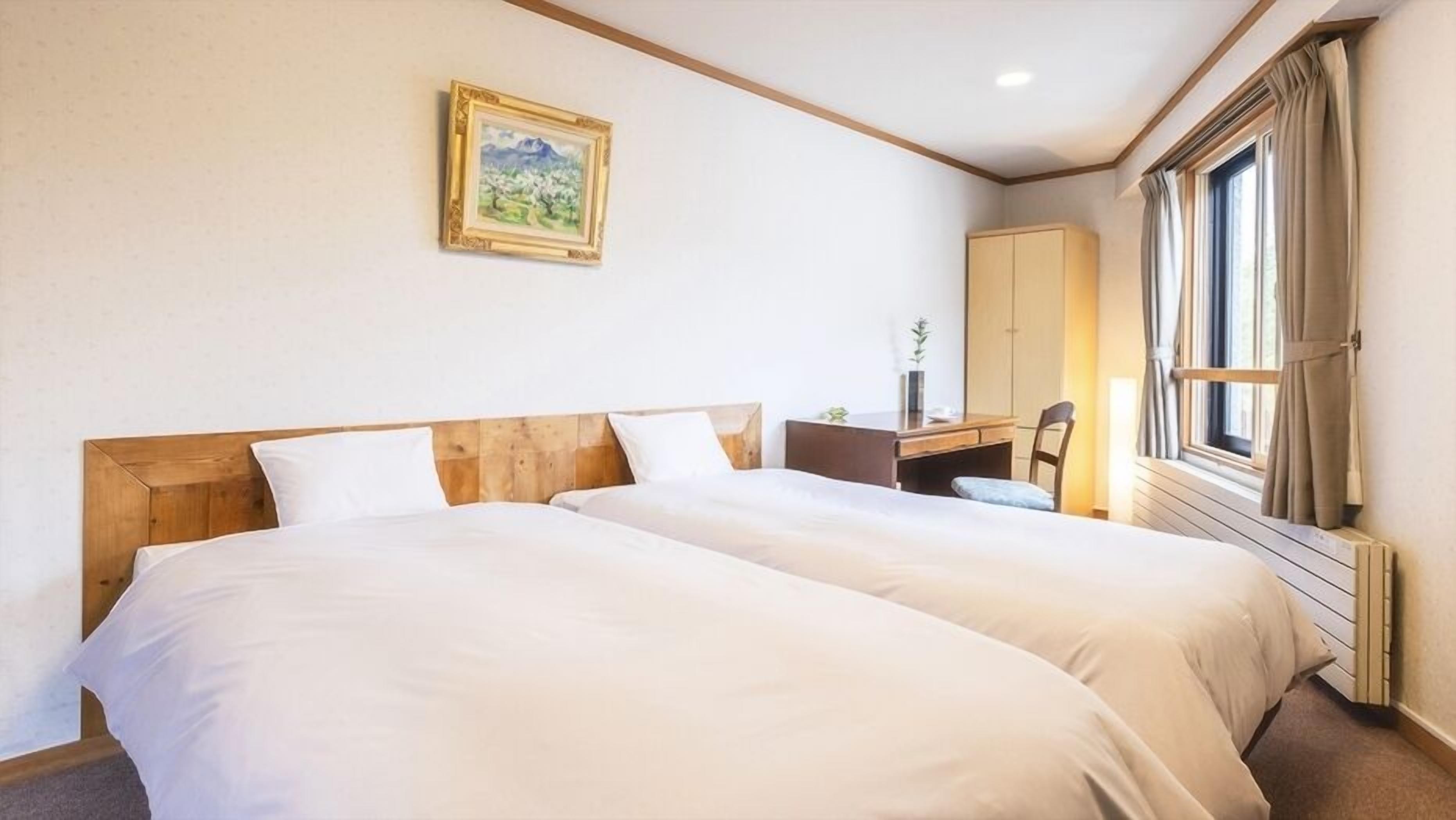 Habitación de hotel con dos camas, un escritorio, una silla, una ventana con cortinas y un cuadro en la pared.
