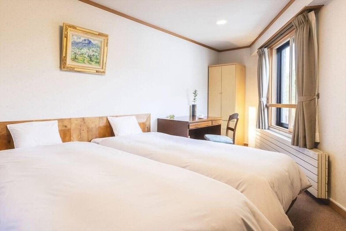 Una camera d'albergo con due letti, una scrivania, una sedia, una finestra con tende e un quadro appeso al muro.