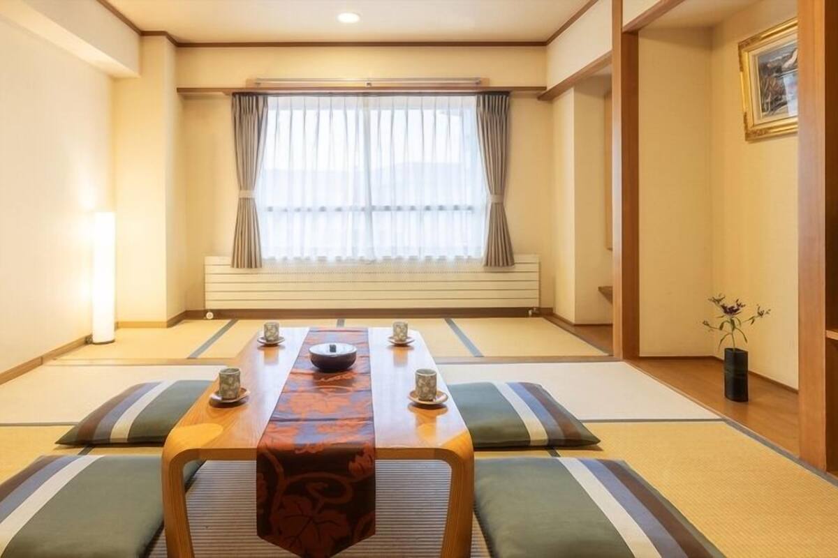 Una camera tradizionale in stile giapponese con pavimento in tatami, un tavolino con runner decorativo, cuscini e uno schermo scorrevole che dà su un'altra stanza.