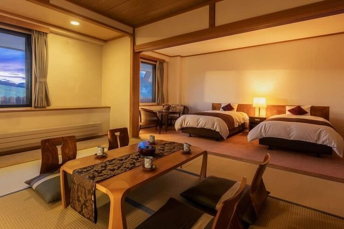 Camera di un tradizionale hotel giapponese, con pavimento in tatami, un tavolino con stuoia intrecciata, due letti con testiere in legno e vista sul paesaggio attraverso ampie finestre.