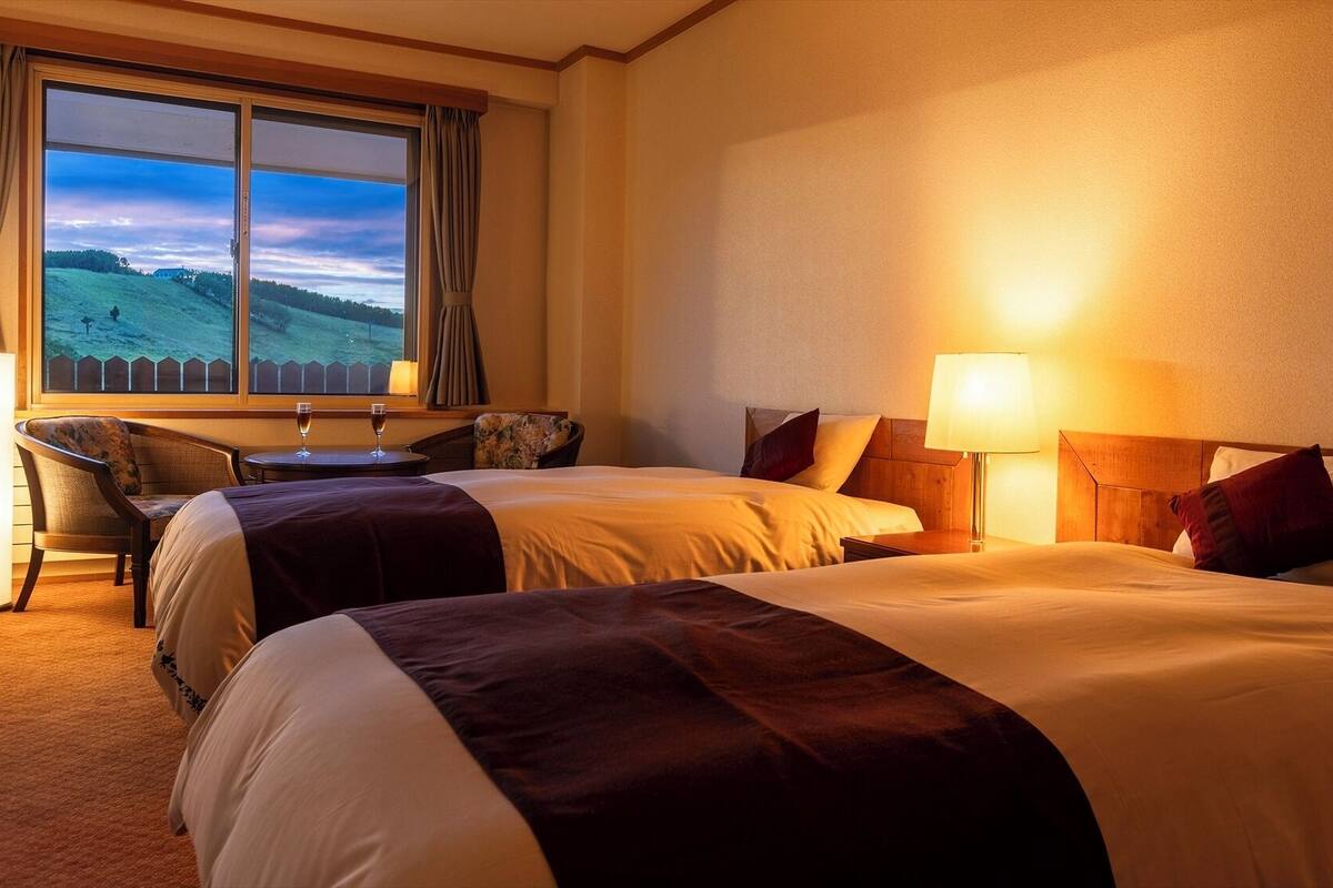 Una camera d'albergo con due letti, una scrivania con sedia, una lampada e vista sul paesaggio dalla finestra.